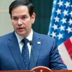 US Troops Suffer: Rubio’s Timeline in Doubt A man in a blue suit speaking into a microphone with an American flag in the background