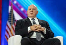 Man in suit sitting on stage with American flag.