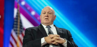 Man in suit sitting on stage with American flag.