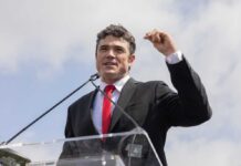 A man in a black suit and red tie speaking at a podium outdoors
