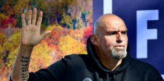Person raising hand while speaking at an outdoor event with autumn foliage in the background
