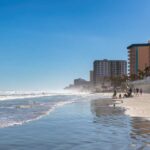 A beach with people walking along the shore and buildings in the background