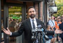 Man in a suit speaking at a press conference with microphones in front of him