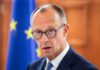 A man in a suit with glasses, looking serious in front of an EU flag
