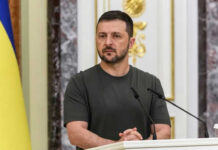 Insider Claims: Zelensky’s Dark Secret Man speaking at podium with microphones, Ukrainian flag behind.