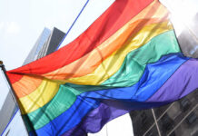 Pride Month Playbook Hits Emergency Alerts A vibrant rainbow flag waving against a city backdrop