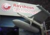 Radar Arms Race: Raytheon’s $100M Gamble Two workers in hard hats and masks review a tablet in a factory