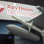 Radar Arms Race: Raytheon’s $100M Gamble Two workers in hard hats and masks review a tablet in a factory