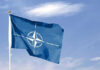 Border Games Rattle NATO—Poland on Alert Blue NATO flag with white emblem under cloudy sky.