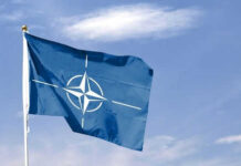 Border Games Rattle NATO—Poland on Alert Blue NATO flag with white emblem under cloudy sky.