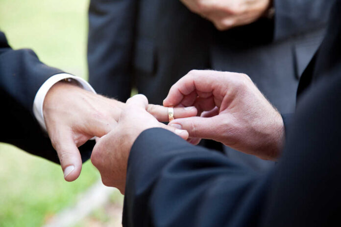 One,Groom,Placing,The,Ring,On,Another,Man's,Finger,During Person wearing rainbow ribbon and heart pin on denim