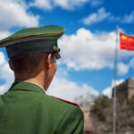 Soldier in a green military uniform facing the Great Wall of China with the Chinese flag in the background