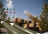 Iran Barrage Tested — Bases Still Standing? Military missiles displayed outdoors with Iranian flags in the background