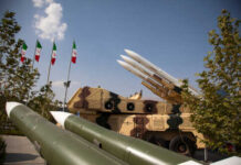 Iran Barrage Tested — Bases Still Standing? Military missiles displayed outdoors with Iranian flags in the background