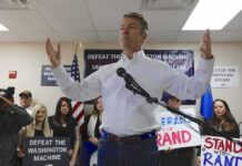 Wuhan Fix? FBI Blocks Airport Grill man in white shirt speaking at a rally with supporters and signs behind him