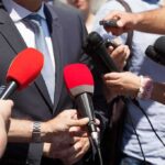 Person in suit surrounded by microphones during media interview