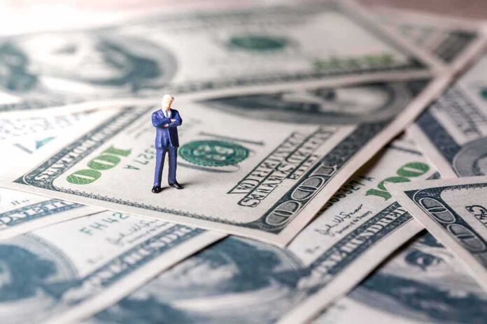shutterstock_1064329202 Miniature businessman standing on US dollar bills