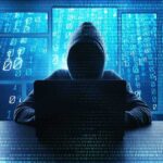 Hooded figure using a laptop in front of glowing code