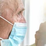 Older man wearing a surgical mask looking out a window