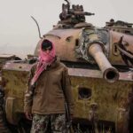 Nationwide Uprising Rumors Rattle Kabul Man in scarf stands before a rusted tank in a barren field