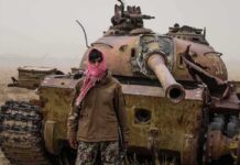 Man in scarf stands before a rusted tank in a barren field