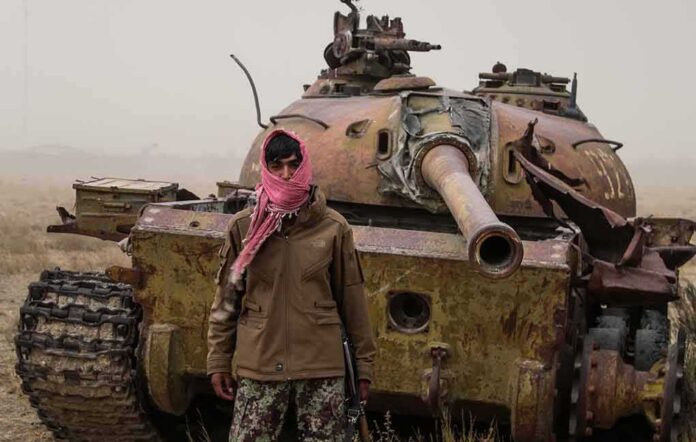 Man in scarf stands before a rusted tank in a barren field