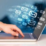 Hand typing on laptop with email icons emerging from screen