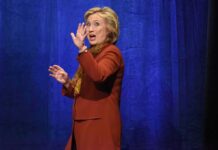 Cathedral Send-Off Teased—Clinton Whispers woman in orange coat gesturing on stage