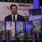 Man speaking at podium on stage, projection behind him.