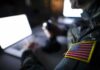 Military Health Heist – Troops Targeted In Bold TRICARE Grift Soldier with U.S. flag patch using a laptop in a dim room