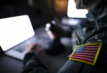 Military Health Heist – Troops Targeted In Bold TRICARE Grift Soldier with U.S. flag patch using a laptop in a dim room