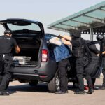 Black Axe Exposed: Romance Racket Busted—Flown In Under Guard customs officers detain a person beside a car with its trunk open