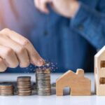 Hand stacking coins beside wooden house models