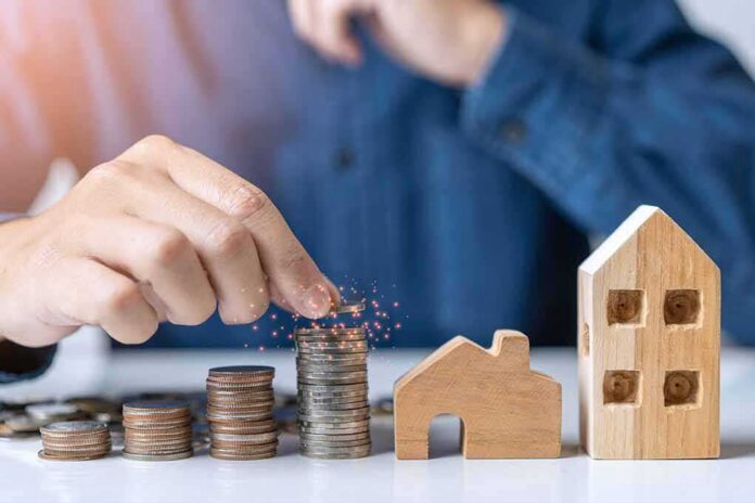 shutterstock_1707356269 Hand stacking coins beside wooden house models