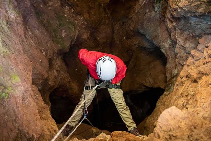 shutterstock_1867616776 Caver in red jacket rappelling into a rocky cave entrance