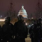 Riot police in protective gear stand before a domed capitol building at night