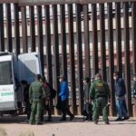 Border agents escort a group near a tall fence and a patrol van