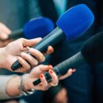 Hard Line Holds: Carrier Slides Into Hormuz Reporters holding colorful microphones
