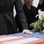 Iran Strike Hits Home—Queens Mourns Mourner rests hand on flag-draped casket with flower arrangement