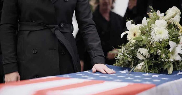 shutterstock_2416028699 Mourner rests hand on flag-draped casket with flower arrangement