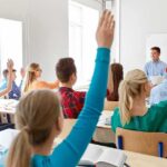 Students raise hands in a classroom with a teacher at the front
