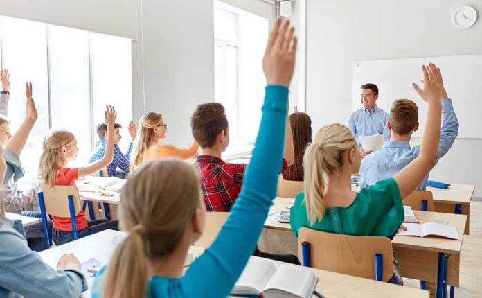 Students raise hands in a classroom with a teacher at the front
